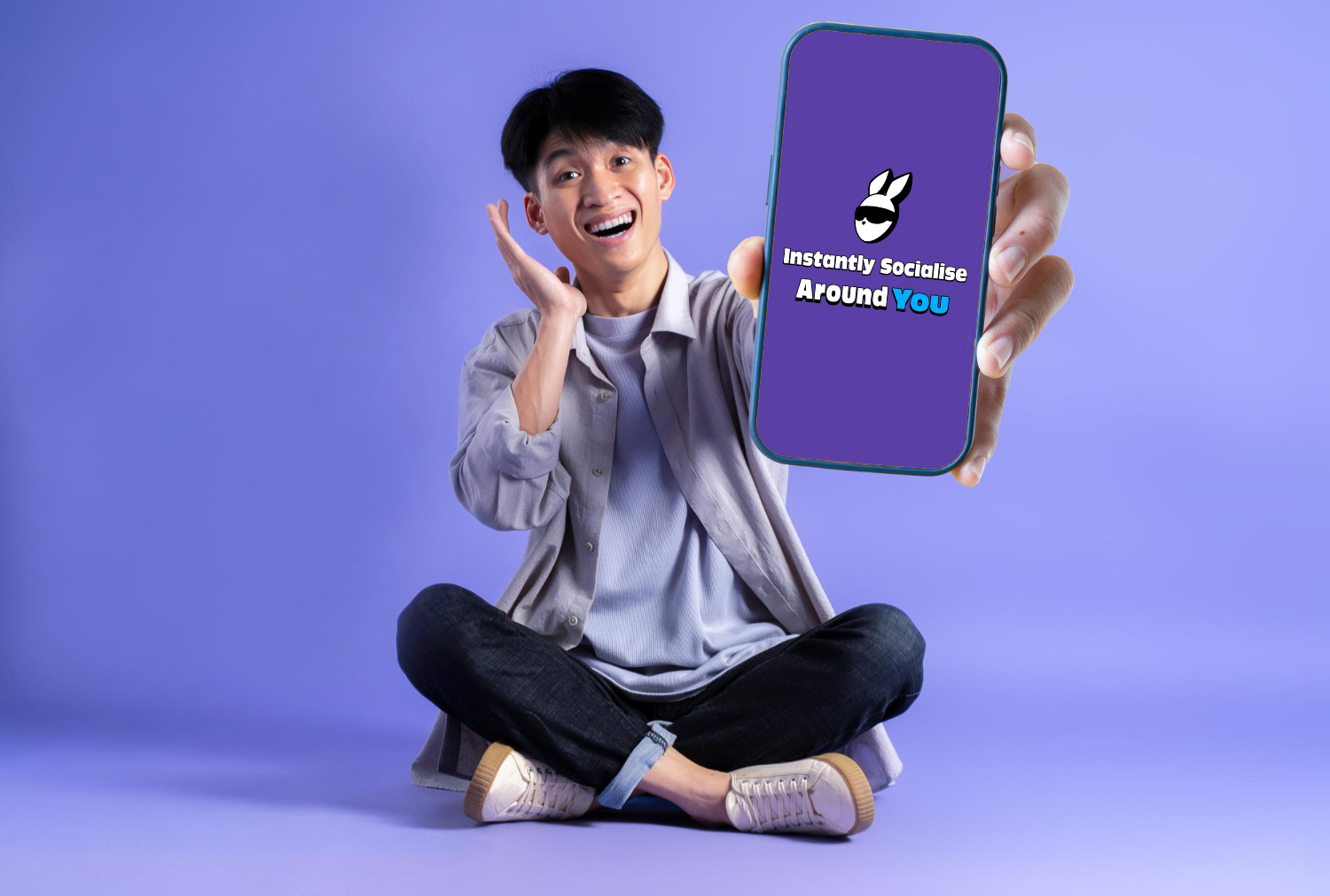 A_young_man_sitting_cross-legged_on_the_floor_enthusiastically_holds_up_a_smartphone_towards_the_camera.The_phone_screen_displays_a_logo_with_the_text"Instantly_Socialise_Around_You"_against_a_purple_background._The_man's_expression_is_excited,_and_the_background_is_a_soft_shade_of_purple,_highlighting_the_vibrant_and_energetic_tone_of_the_image,_which_promotes_social_connection_via_technology.