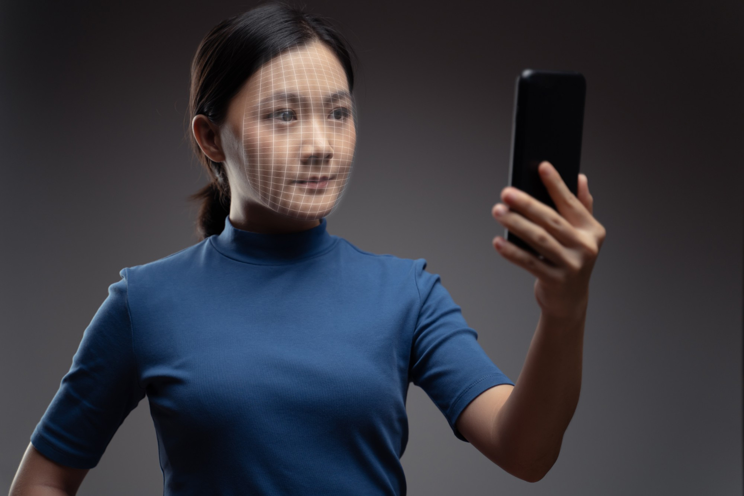 A_woman_holds_up_a_smartphone_while_a_digital_grid_scans_her_face,_indicating_the_use_of_facial_recognition_technology._She_appears_calm_and_focused,_and_the_background_is_neutral,_allowing_the_focus_to_remain_on_the_face-scanning_process._The_image_highlights_the_advancement_of_biometric_security_and_identity_verification_through_modern_technology.