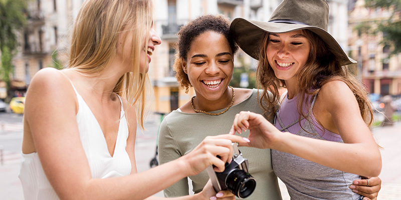 Three_female_friends_stand_together_outdoors,_smiling_and_laughing_as_they_look_at_photos_on_a_camera._One_of_them,_wearing_a_sun_hat,_appears_excited_as_she_points_at_the_screen._They_are_in_a_city_setting,_surrounded_by_buildings,_and_the_moment_captures_their_joy_and_bond_while_enjoying_time_together