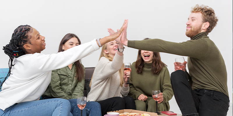 A_group_of_friends_sits_on_a_couch,_sharing_laughs_and_drinks,_while_two_of_them_high-five_each_other_enthusiastically._A_table_in_front_of_them_is_set_with_a_pizza_and_drinks,_adding_to_the_casual_and_friendly_gathering._The_mood_is_joyful_and_celebratory_as_the_group_enjoys_their_time_together.