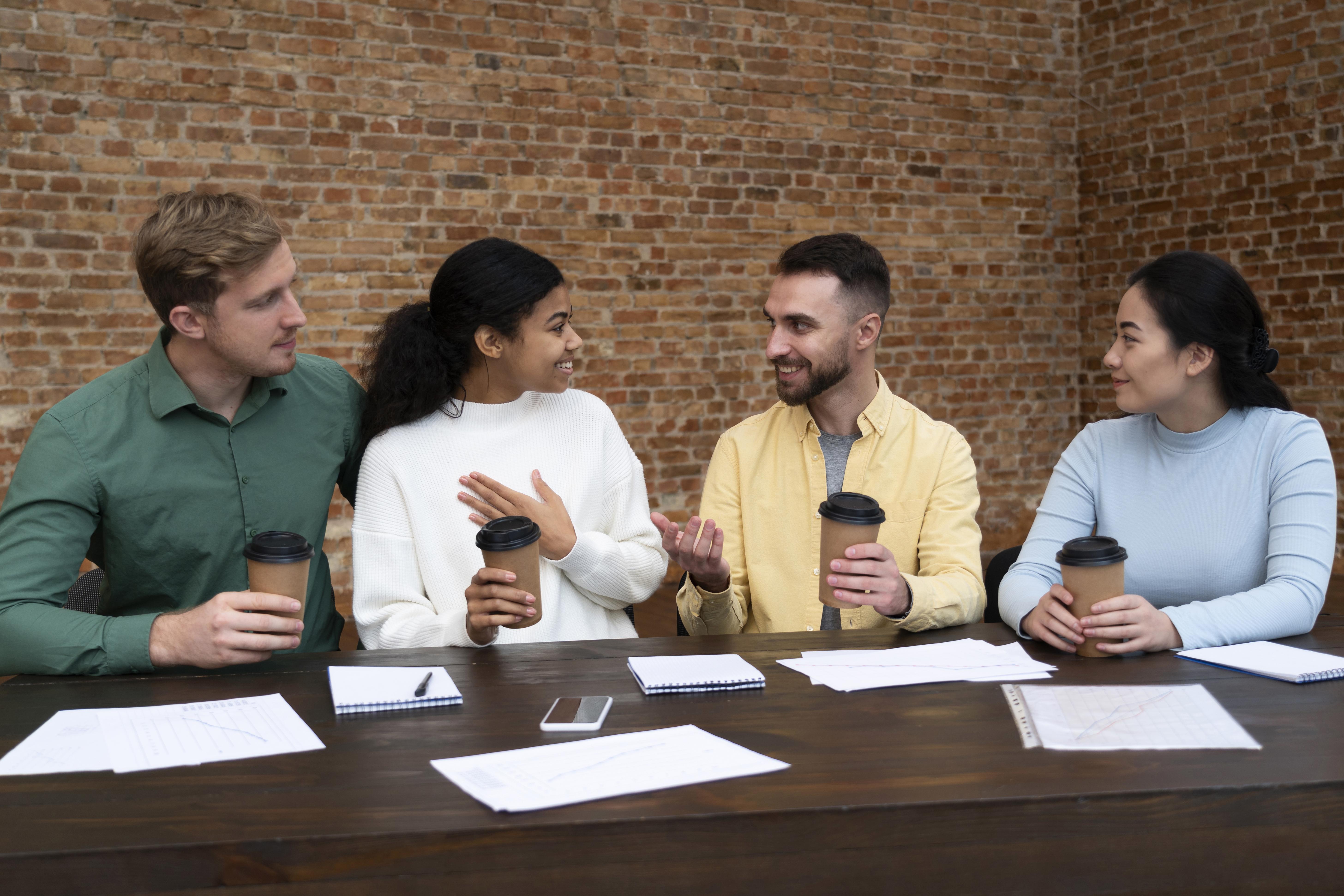 A_group_of_four_people_sits_at_a_table_in_a_casual_work_environment,_engaged_in_a_lively_conversation_while_holding_coffee_cups._Papers_and_notebooks_are_spread_out_on_the_table,_indicating_a_brainstorming_or_collaborative_meeting._The_brick_wall_background_and_relaxed_body_language_suggest_an_informal_but_productive_discussion.