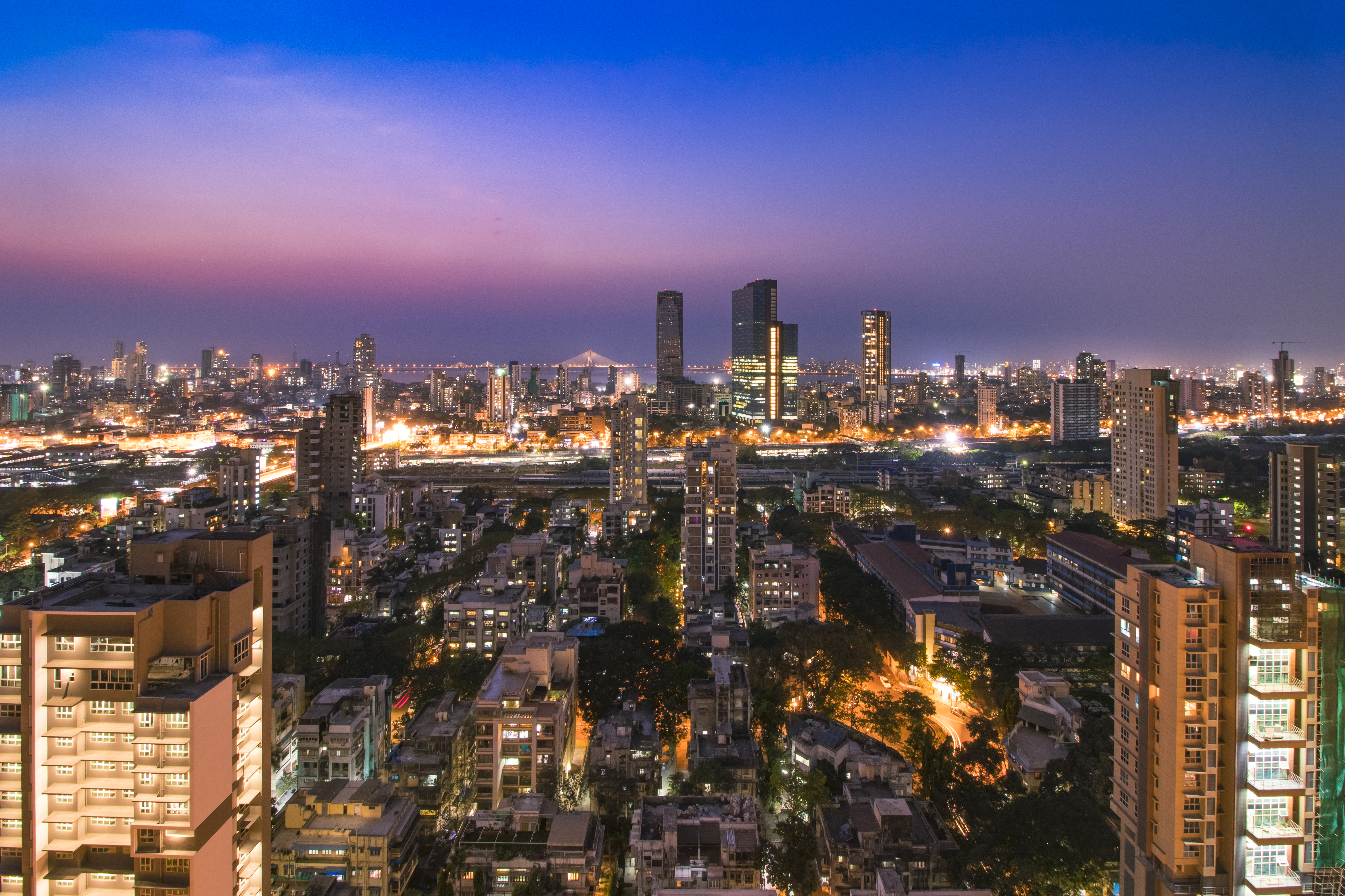 A_vibrant_cityscape_of_Mumbai_at_dusk,_showing_a_sprawling_skyline_with_tall_buildings_lit_up_against_a_gradient_sky_of_blue_and_purple._In_the_distance,_the_Bandra-Worli_Sea_Link_bridge_is_visible,_connecting_the_city_across_the_water._The_streets_and_buildings_below_are_aglow_with_lights,_highlighting_the_bustling_energy_of_the_urban_landscape_as_night_falls.