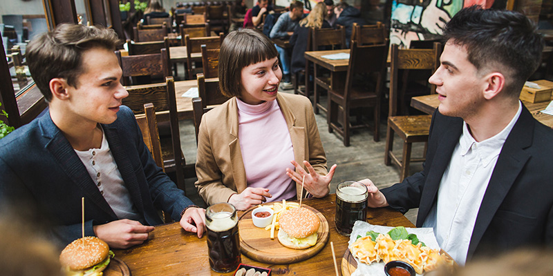 Three_friends_sit_together_at_a_restaurant_table,_engaged_in_conversation_while_enjoying_burgers,_fries,_and_drinks._The_setting_is_warm_and_inviting,_with_the_friends_smiling_and_talking_animatedly._The_meal_and_drinks_add_to_the_casual_and_friendly_atmosphere_of_their_gathering