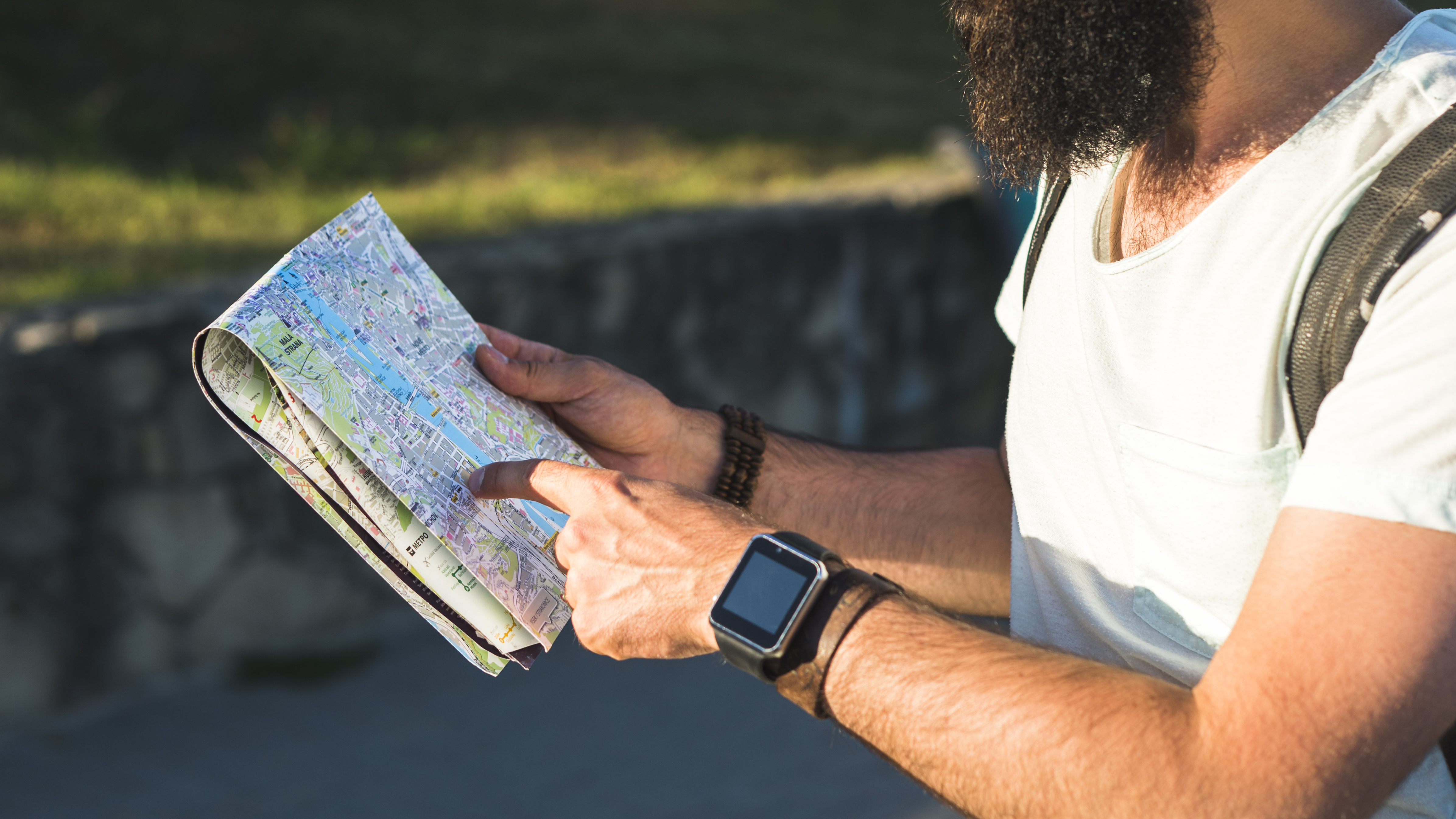 A_man_with_a_beard_and_wearing_a_smartwatch_holds_a_folded_map,_pointing_at_a_location_as_he_explores_an_outdoor_area._He_is_dressed_casually_in_a_t-shirt_with_a_backpack_on_his_shoulder,_suggesting_he_is_on_a_trip_or_hiking._The_scene_is_set_against_a_blurred_background_of_nature_or_a_stone_wall,_capturing_a_moment_of_navigation_during_a_vacation_or_adventure.