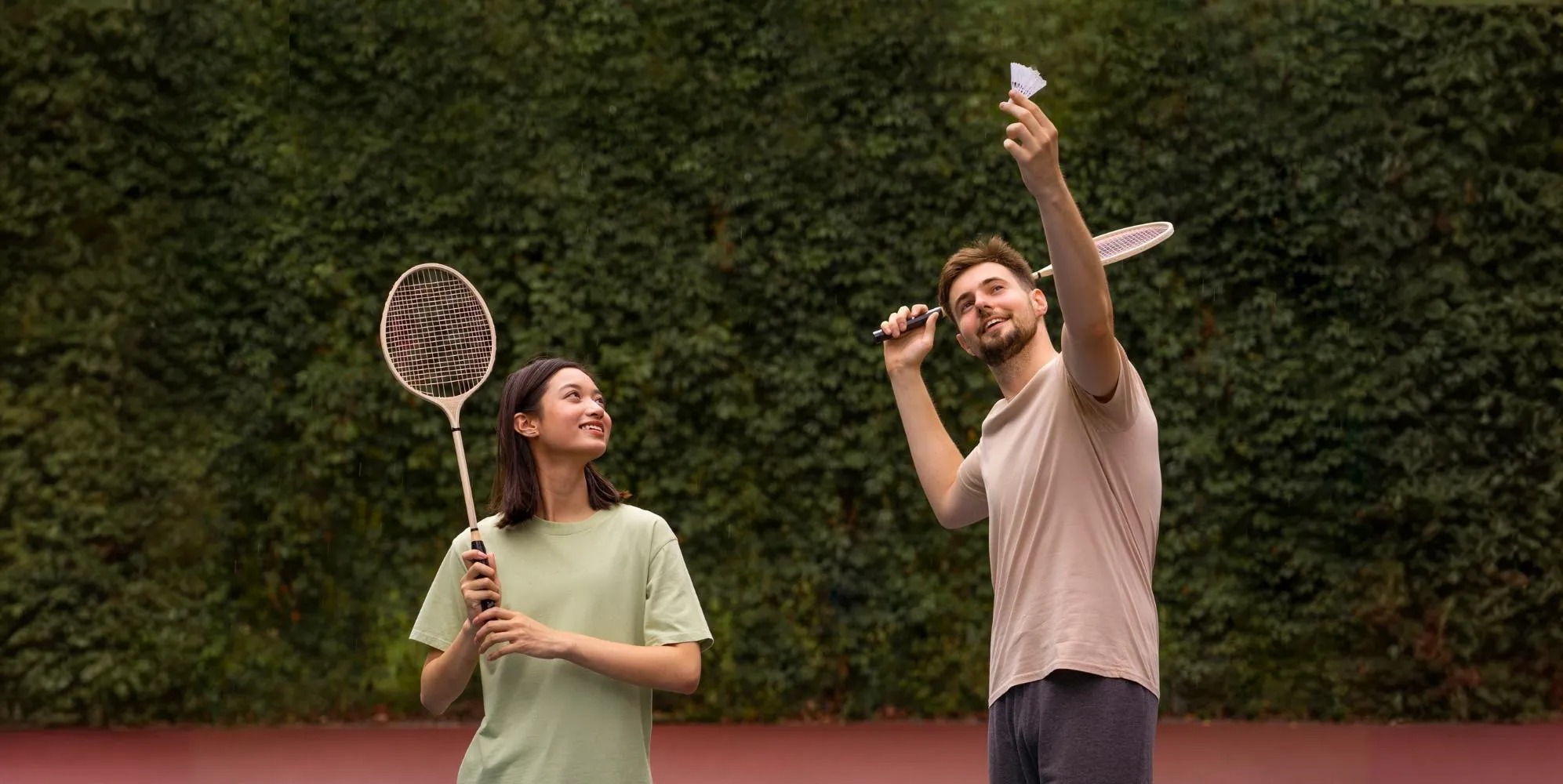 A_man_and_a_woman_are_playing_badminton_outdoors,_both_holding_rackets_and_looking_upward_as_the_man_prepares_to_serve_the_shuttlecock._They_are_dressed_in_casual_clothes,_standing_on_a_court_surrounded_by_greenery,_and_their_expressions_suggest_they_are_enjoying_the_game_in_a_relaxed_and_fun_atmosphere.