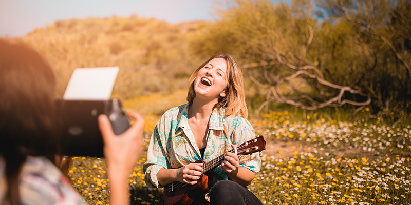 A_woman_is_sitting_in_a_field_of_wildflowers,_playing_a_ukulele_and_laughing_joyfully_while_her_friend_takes_a_photo_of_her._The_sun_is_shining_brightly,_and_the_background_shows_bushes_and_a_clear_sky,_creating_a_vibrant_and_cheerful_scene_of_friends_capturing_a_moment_of_happiness_in_nature.