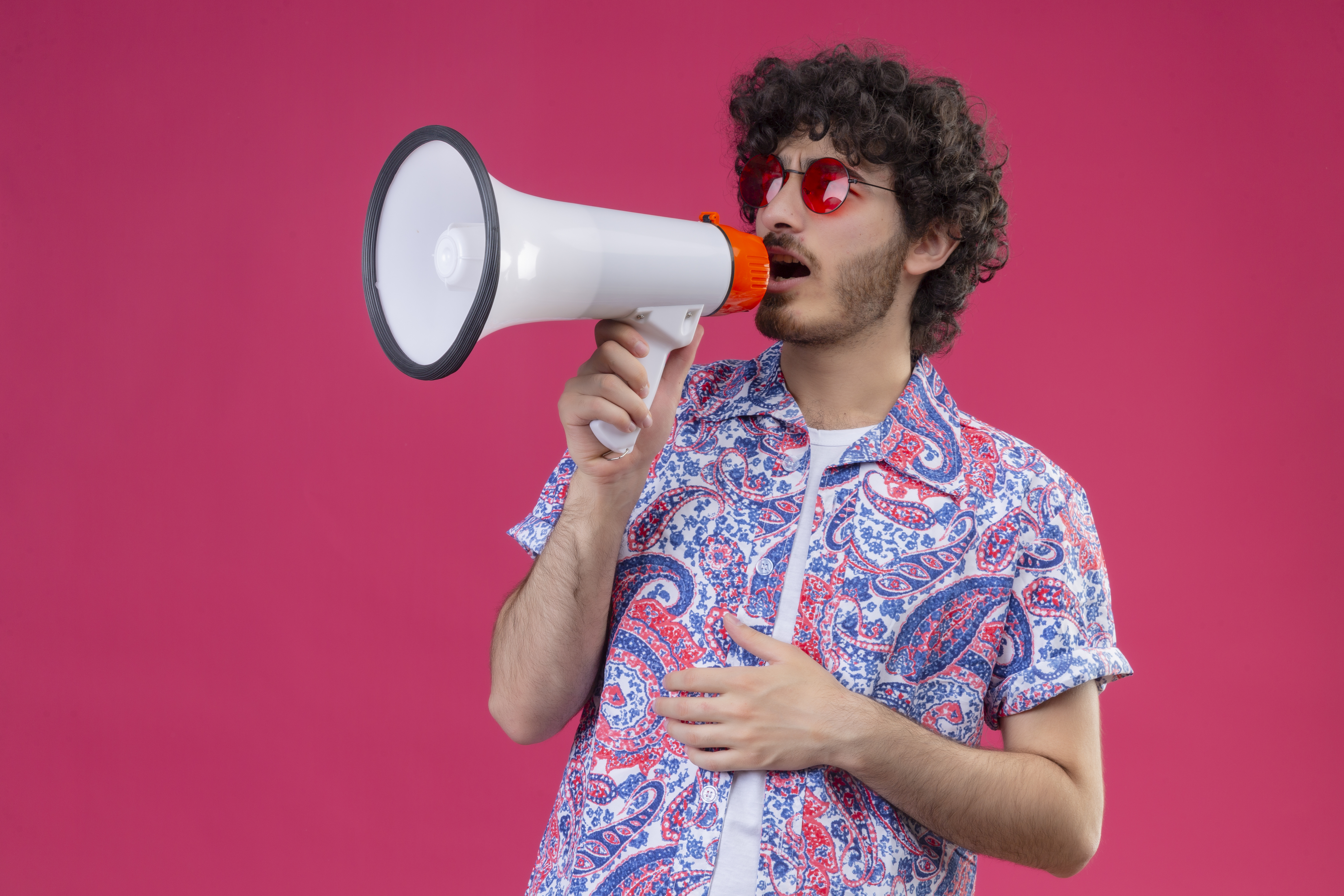 A_young_man_with_curly_hair_and_wearing_red_sunglasses_holds_a_megaphone_while_speaking,_with_one_hand_on_his_belly._He_is_dressed_in_a_colorful_paisley_shirt,_and_the_background_is_a_bright_pink_wall._The_image_conveys_a_vibrant_and_expressive_mood,_highlighting_his_loud_announcement_or_statement.